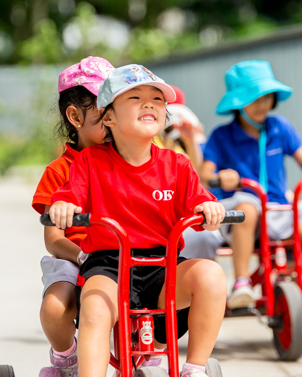 A group of female OFS Kindergarten students riding tricycles during recess in the Kindergarten playground