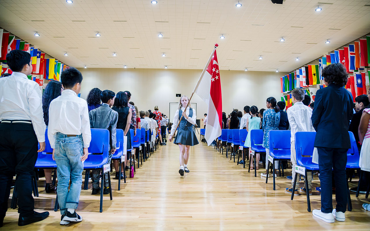 A female OFS Elementary School student carrying Singapore flag walking towards the stage in auditorium while other students stand up during Elementary School Model United Nations (PY-MUNOFS)